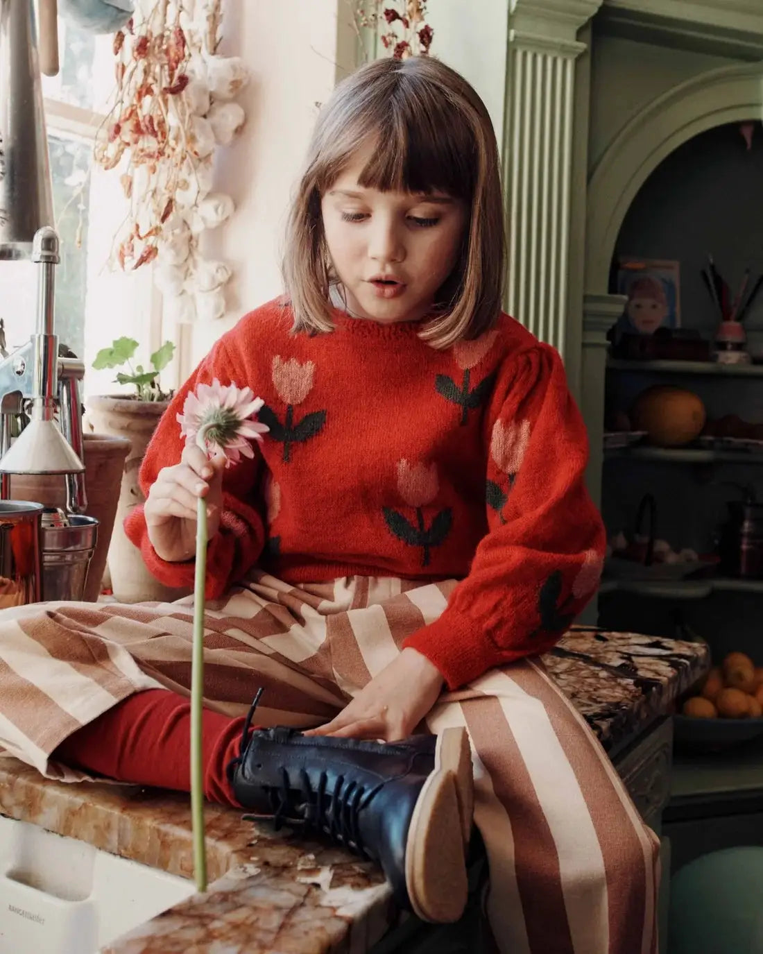 EMILE ET IDA pull intarsia tulipe mars red sweater with tulip motif; girl sits on a kitchen counter.