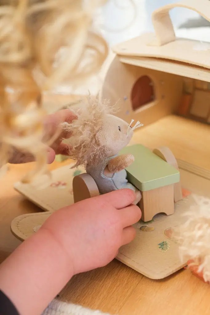 Maison en rondins pour poupées, bois naturel; enfant joue près d’une peluche sur un meuble en bois.