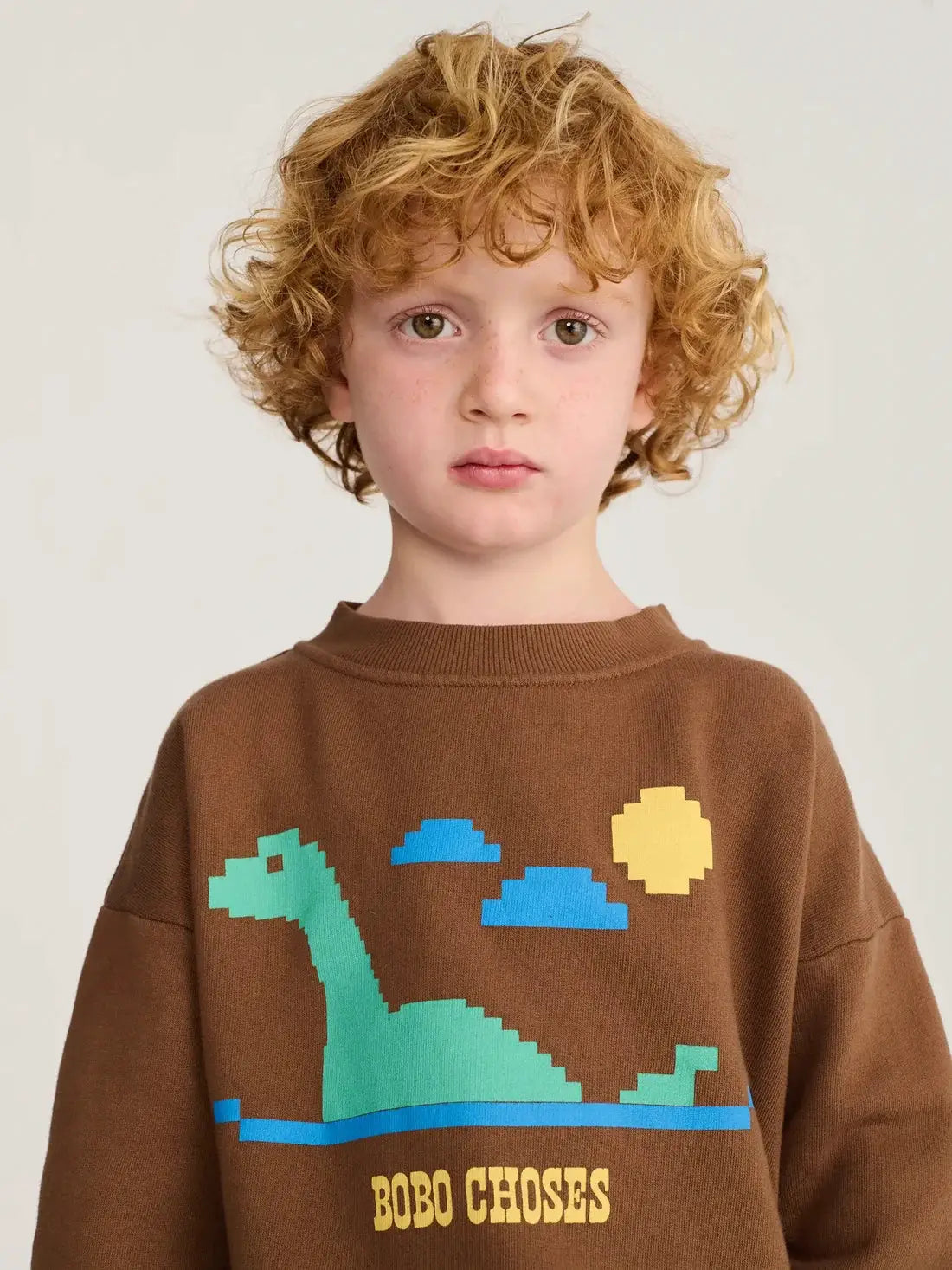 BOBO CHOSES Lake Monster sweatshirt, brown with a pixel green dinosaur graphic and blue water.