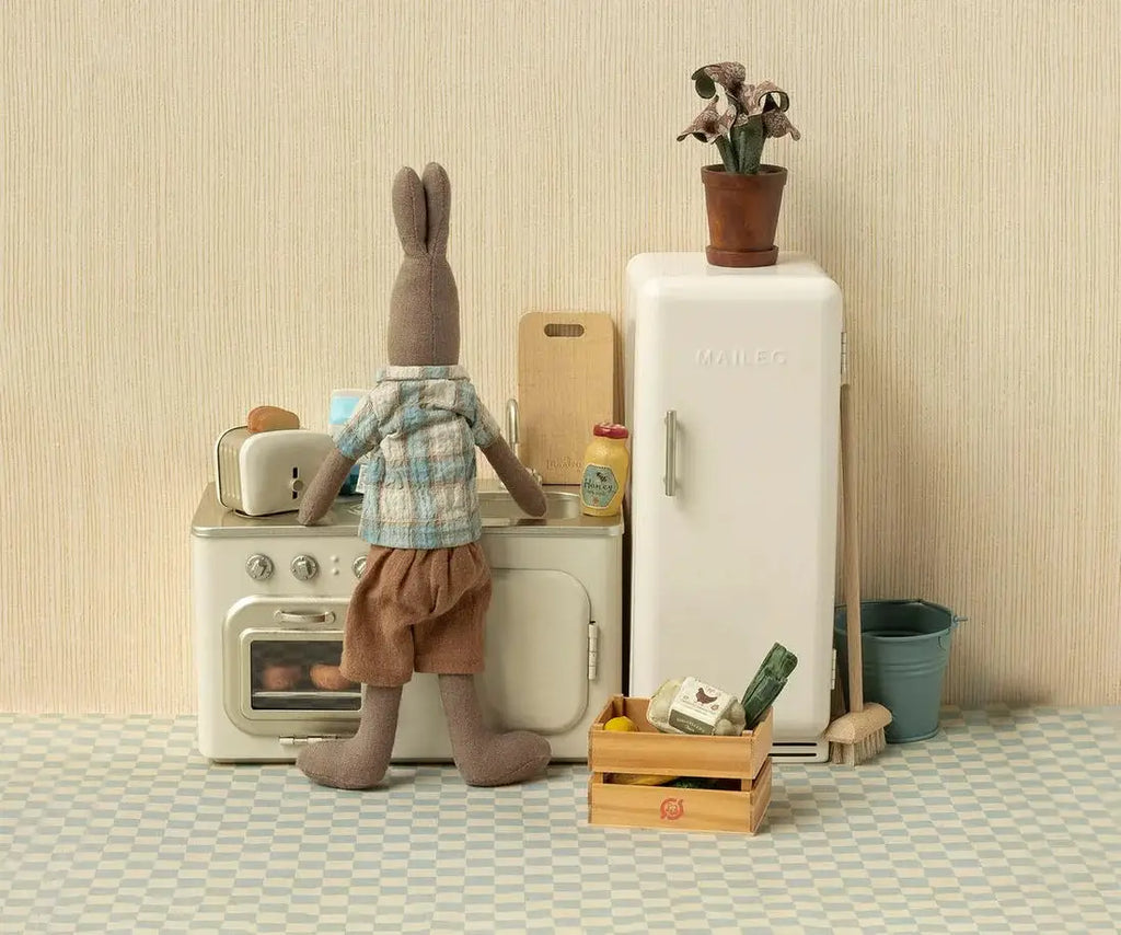 Maileg canapé miniature kitchen scene; rabbit figure at stove beside white fridge with potted plant.