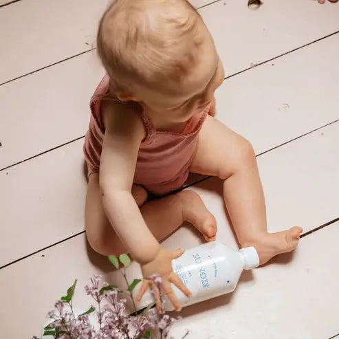 MINOIS mousse de bain bubble bath bottle held by baby on pale wooden floor, white bottle with blue label