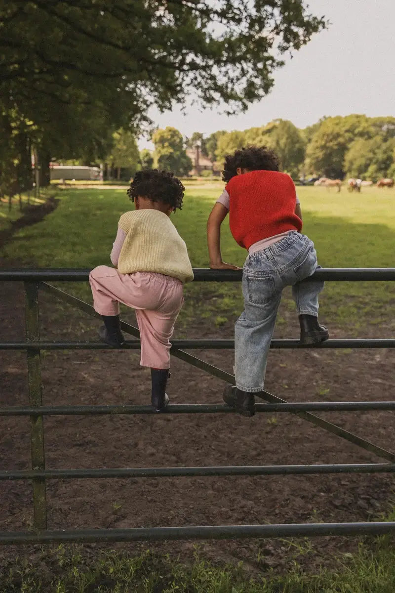 Kids knit pullovers worn by two children on a park fence, one red pullover and one cream vest, with denim jeans.