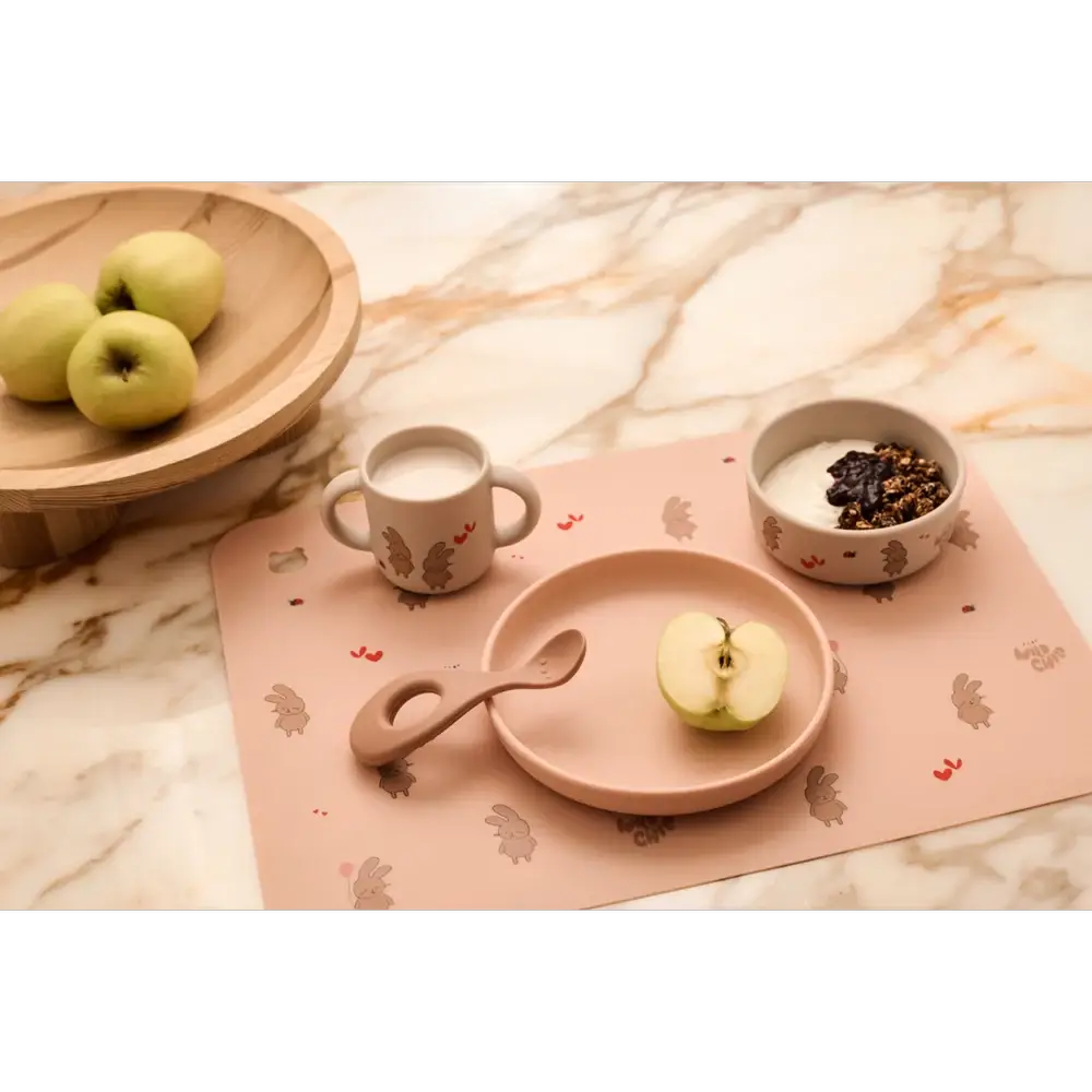 LIEWOOD set repas VIVI rosey pink feeding set (plate, cup, bowl, spoon) on bunny-pattern mat with apples nearby.