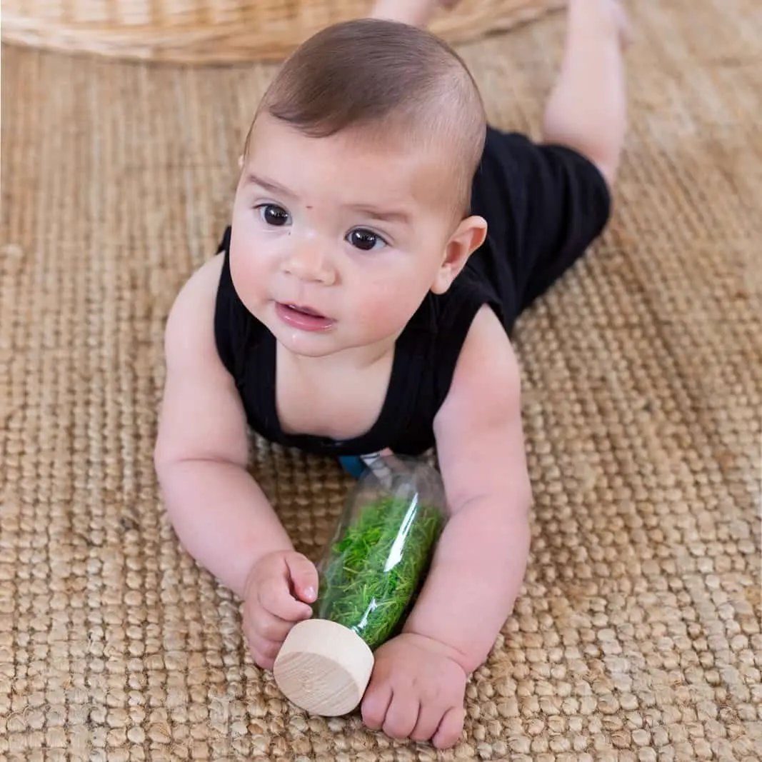 Petit Boum sensory bottle baby toy with wooden cap; clear bottle with green filler, held by a baby on a woven rug