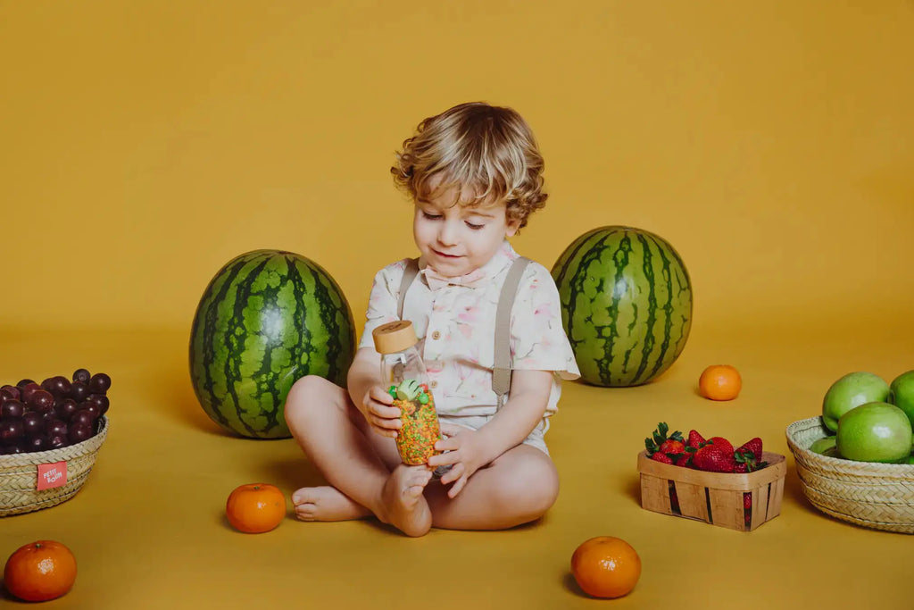 Bouteille sensorielle Petit Boum tenue par un enfant, entourée de fruits: pastèques, oranges et raisins.