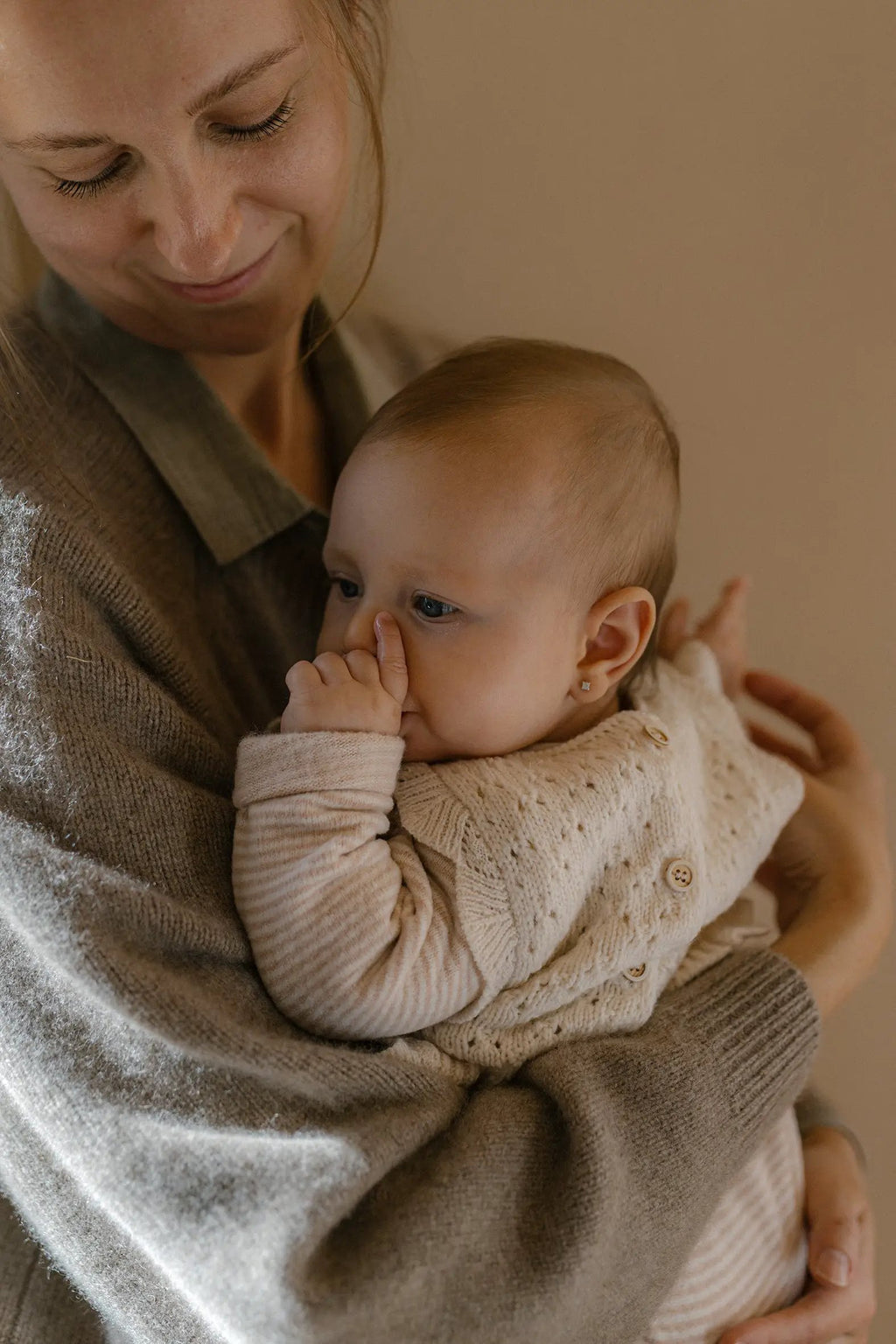 1+ IN THE FAMILY Metz nude baby pyjama by Citron Grenadine; infant in cream knit cardigan held by caregiver