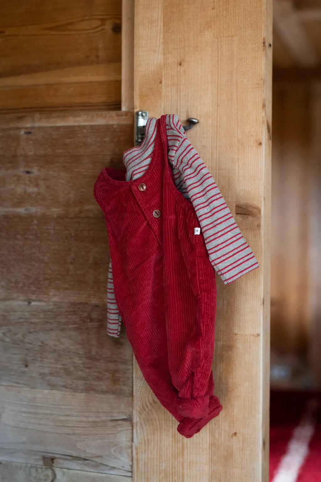 Red corduroy romper layered over a grey and red striped tee, hanging on a wooden post