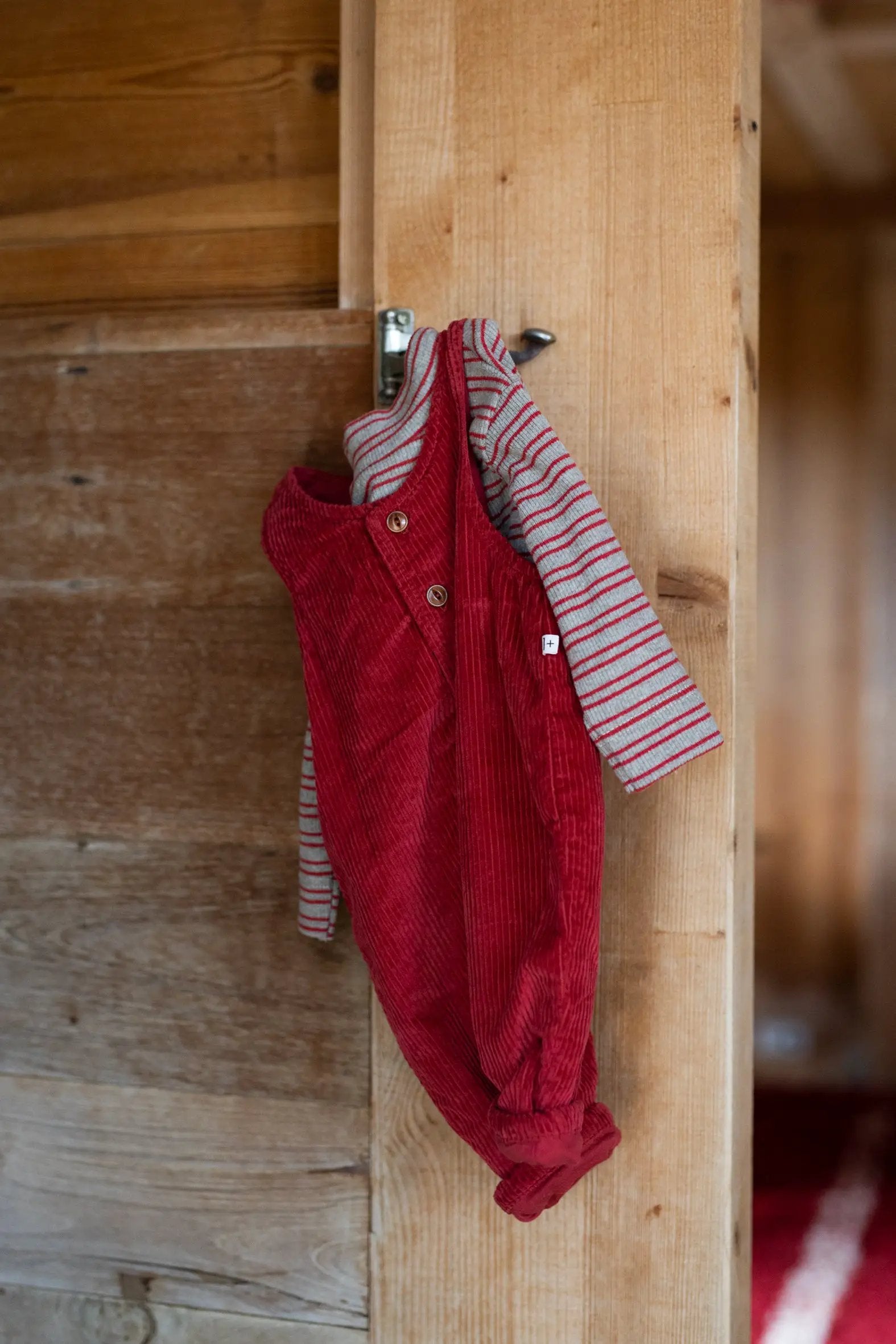 Red corduroy romper layered over a grey and red striped tee, hanging on a wooden post